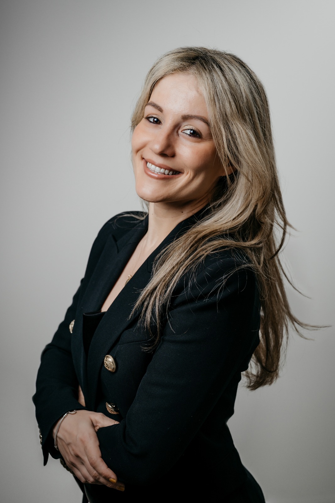 A studio portrait of a woman with long, blonde-highlighted hair smiling directly at the camera against a plain gray background. She is wearing a dark blazer with gold buttons, standing turned slightly to the side with her arms crossed.
