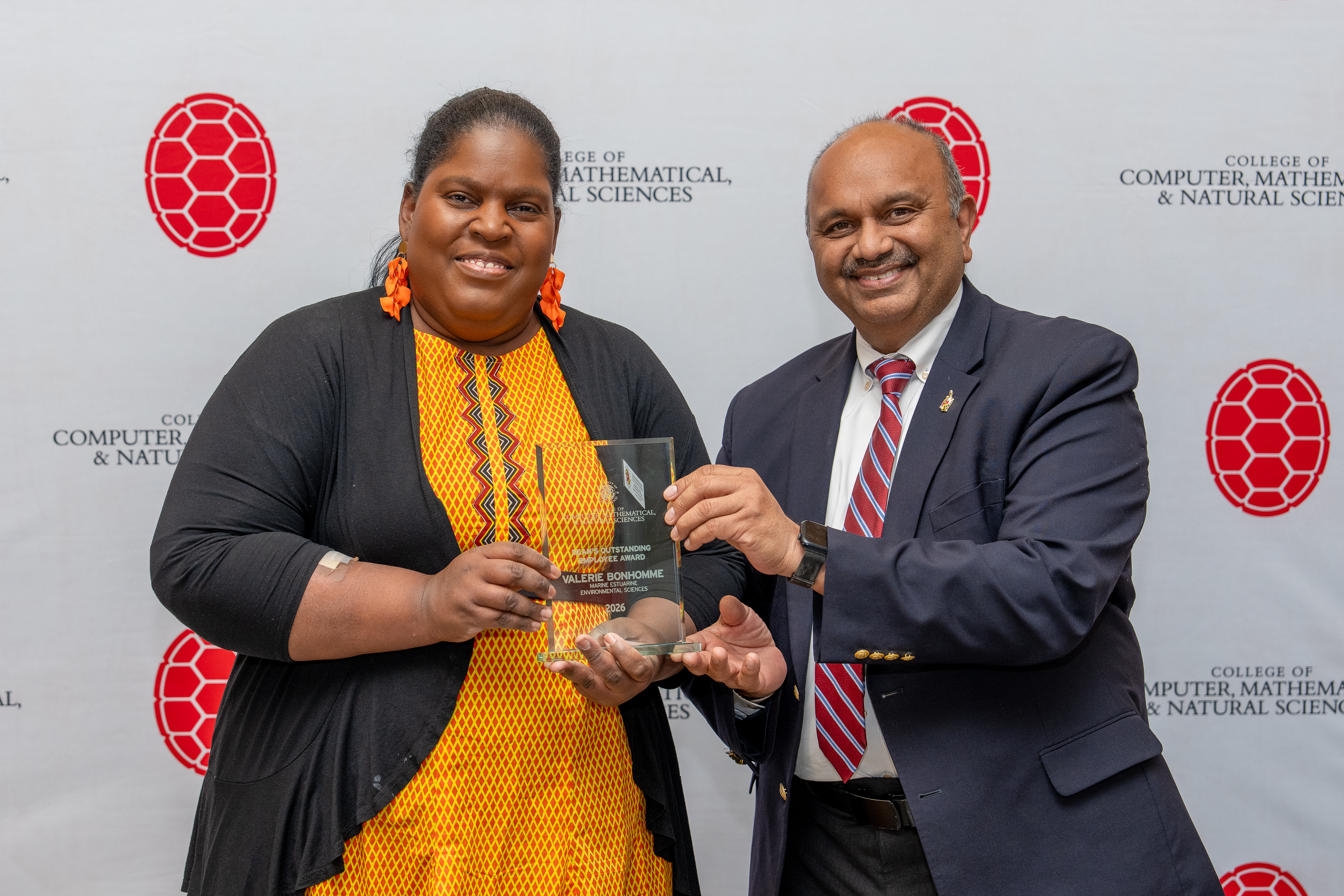 A woman in a black and yellow dress accepts a glass award from a man in a blue suit with a white shirt and red tie. They stand in front of a backdrop with red turtle shells and text saying "College of Computer, Mathematical, & Natural Sciences."