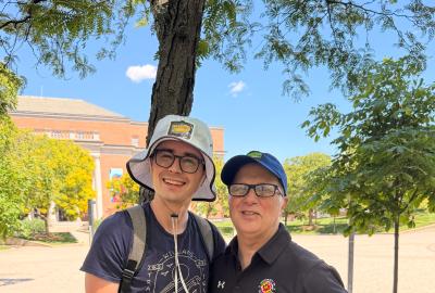 Two men stand outside, smiling on a sunny day. The one on the left wears gray shorts, a navy t-shirt, and a light gray bucket hat. The one on the right wears black jeans, a black University of Maryland promo, and a blue baseball cap.