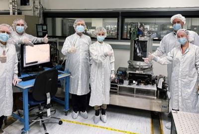 Six people in white coveralls, surgical masks, hairnets, and shoe covers standing in a laboratory setting. They are giving a thumbs-up gesture surrounded by a computer monitor displaying graph data on the left and a large, cylindrical laboratory apparatus on the right.