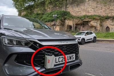 Close-up of a dark grey BYD vehicle parked on a road next to an old stone wall structure. Encircled in red on the front bumper is a small grey plastic box with a red "2" written on it, secured to the front grill next to the UK license plate.