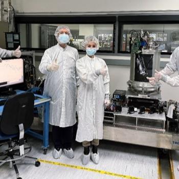Six people in white coveralls, surgical masks, hairnets, and shoe covers standing in a laboratory setting. They are giving a thumbs-up gesture surrounded by a computer monitor displaying graph data on the left and a large, cylindrical laboratory apparatus on the right.