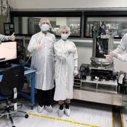 Six people in white coveralls, surgical masks, hairnets, and shoe covers standing in a laboratory setting. They are giving a thumbs-up gesture surrounded by a computer monitor displaying graph data on the left and a large, cylindrical laboratory apparatus on the right.