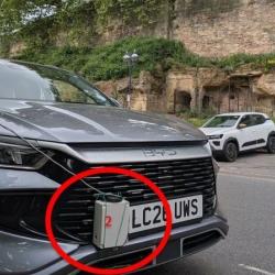 Close-up of a dark grey BYD vehicle parked on a road next to an old stone wall structure. Encircled in red on the front bumper is a small grey plastic box with a red "2" written on it, secured to the front grill next to the UK license plate.