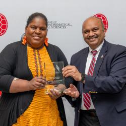 A woman in a black and yellow dress accepts a glass award from a man in a blue suit with a white shirt and red tie. They stand in front of a backdrop with red turtle shells and text saying "College of Computer, Mathematical, & Natural Sciences."